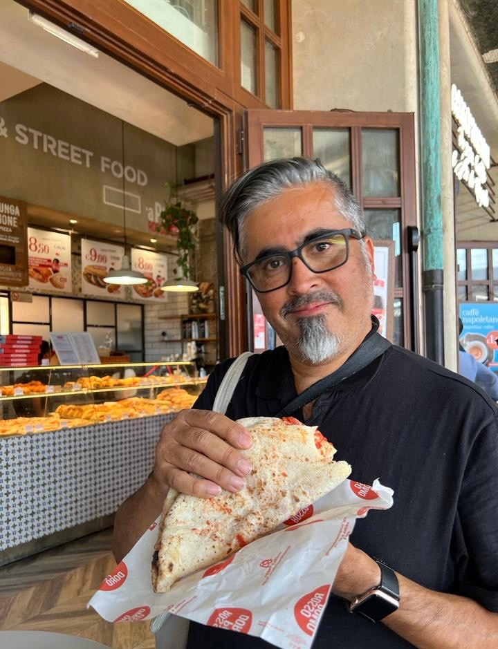 Gene Rocha smiling while holding a delicious pizza against the backdrop of the train station in Florence, Italy.